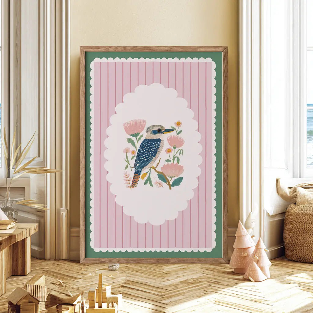 Framed artwork with a bird and floral design on a pink background in a room with wooden floor and decor.
